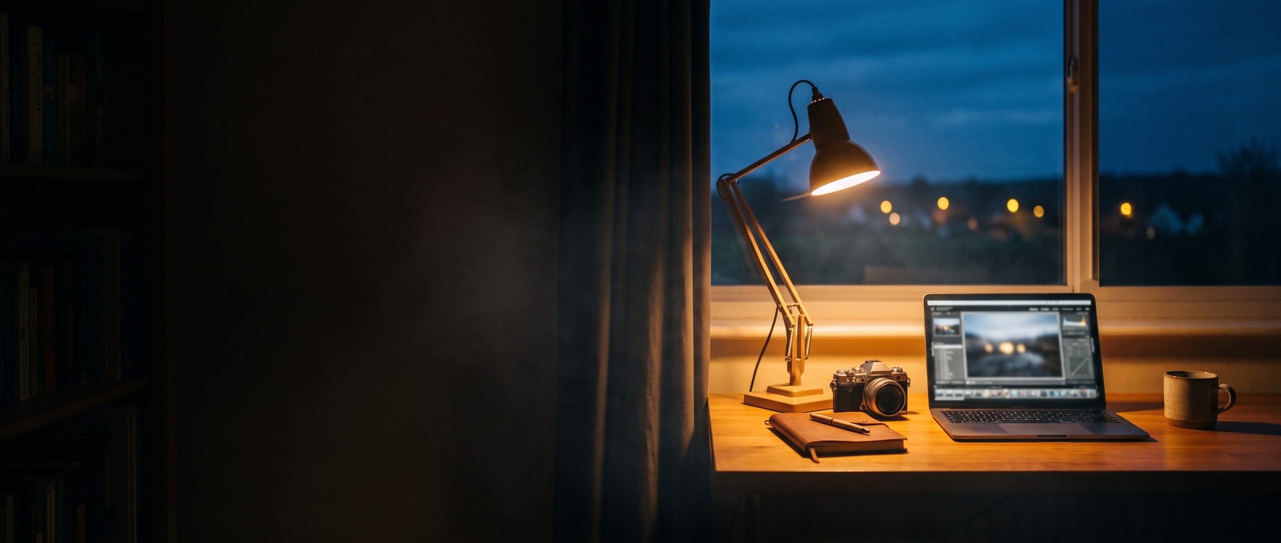 Schreibtisch mit Kamera und Notizbuch im Licht einer Schreibtischlampe, dahinter die blaue Abenddämmerung